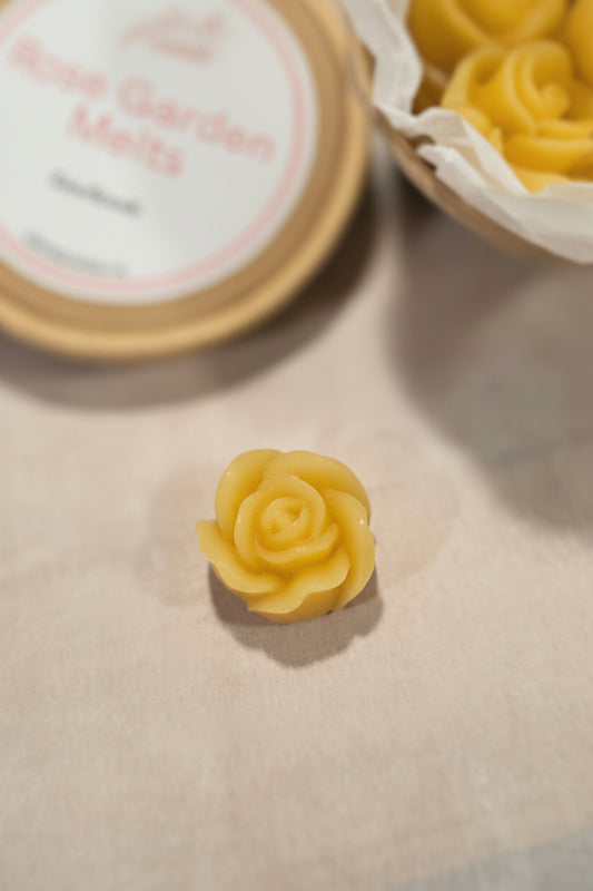 Yellow rose-shaped melts next to a container labeled 'Rose Garden Melts' on a textured surface.